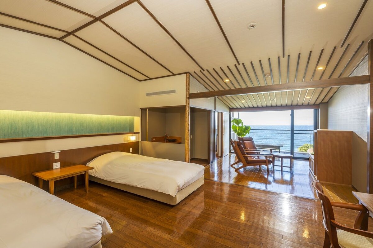 Quarto de hotel com duas camas, chão de madeira, vista para o mar e varanda com assentos.
