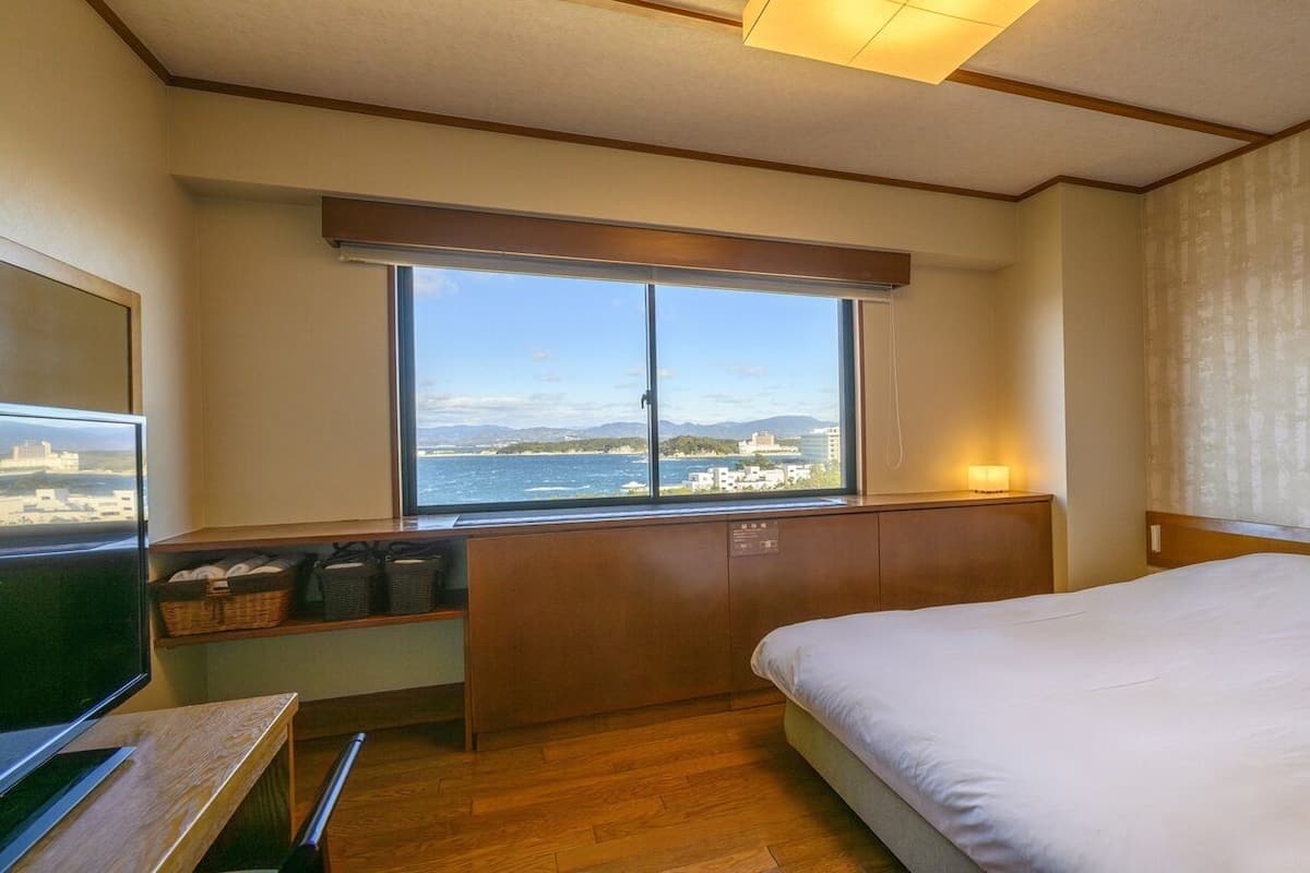 Een hotelkamer met een bed, een bureau, een raam met uitzicht op zee en een nachtkastje.