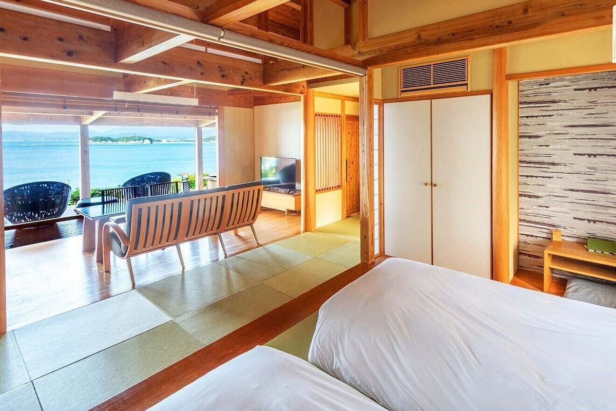 Een kamer met een bed, een televisie, een houten bankje en uitzicht op de zee.
