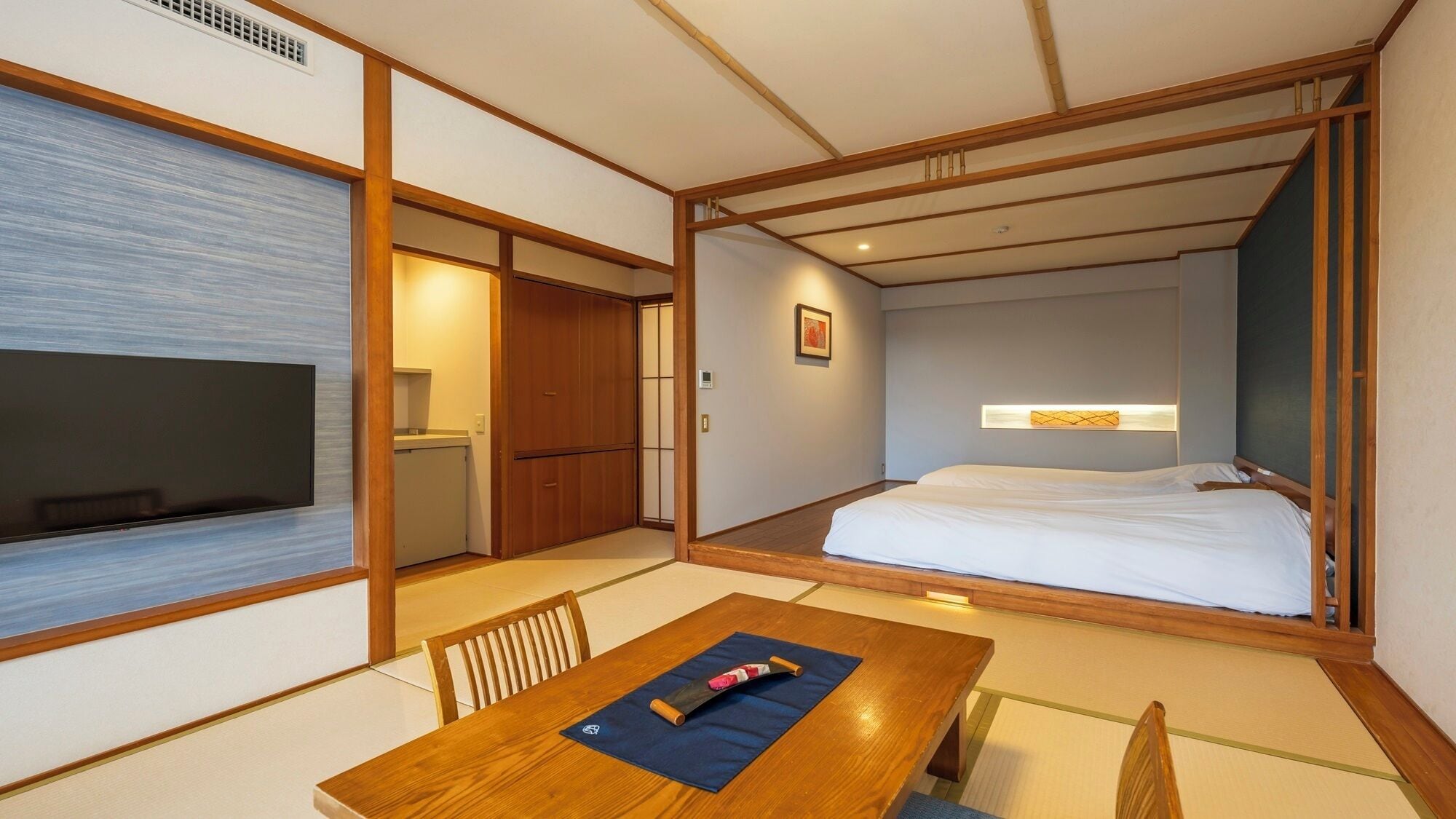 Ein traditioneller japanischer Raum mit Tatami-Boden, einem Holztisch mit Kissen, einem Bett mit weißer Bettwäsche und einem an der Wand montierten Fernseher.