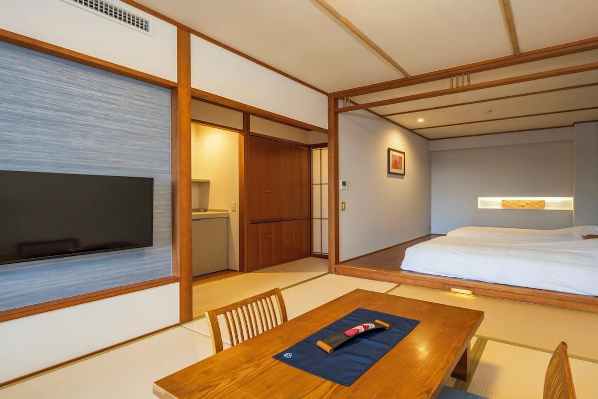 Een traditionele Japanse kamer met tatami vloeren, een houten tafel met kussen, een bed met witte beddengoed en een televisie aan de muur.