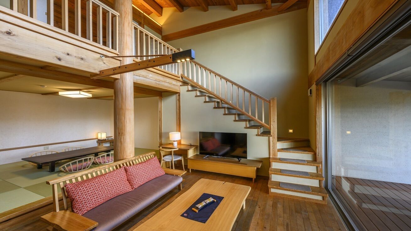 Eine Holztreppe führt zu einer erhöhten Ebene, ein Sofa mit Kissen, ein Holztisch und ein Fernseher in einem Raum mit Holzbalken und einem großen Fenster.