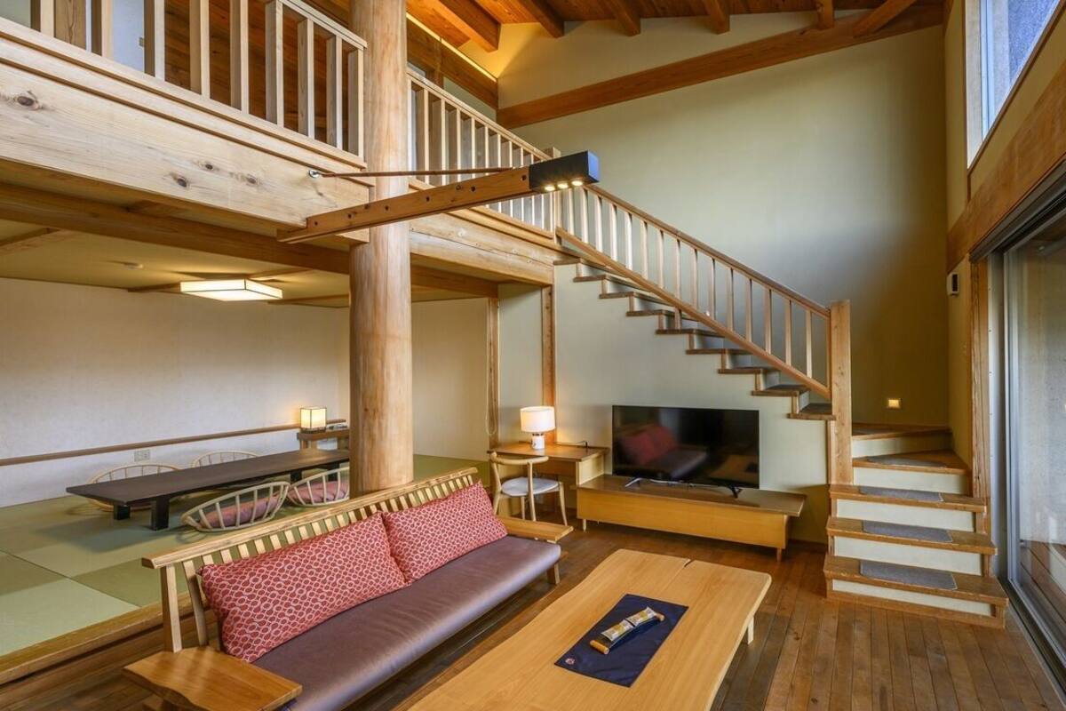 Een houten trap leidt naar een hoger niveau, een bank met kussens, een houten tafel en een televisie in een kamer met houten balken en een groot raam.
