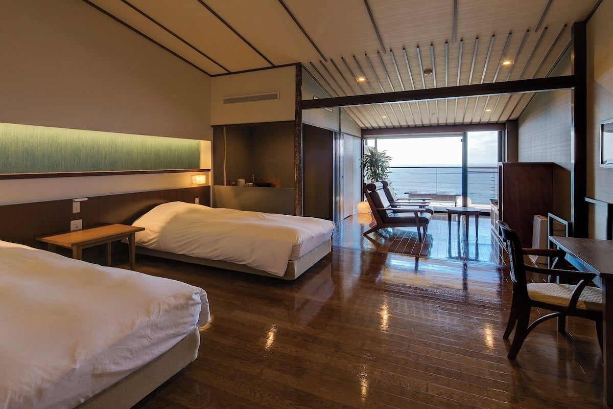 Een hotelkamer met twee bedden, een kleine tafel, een stoel en uitzicht op de oceaan.