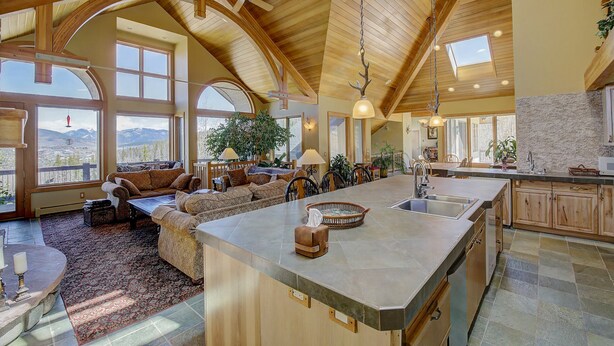 A spacious living area with a large island, wooden ceiling beams, and a view of mountains through large windows.
