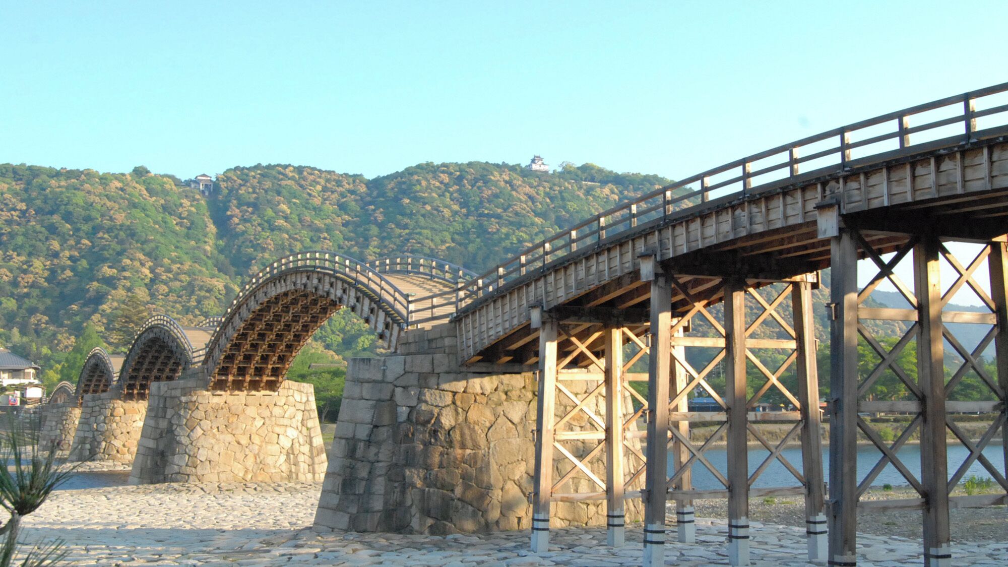 A stone bridge with a wooden walkway, set against a backdrop of green hills and a clear sky.