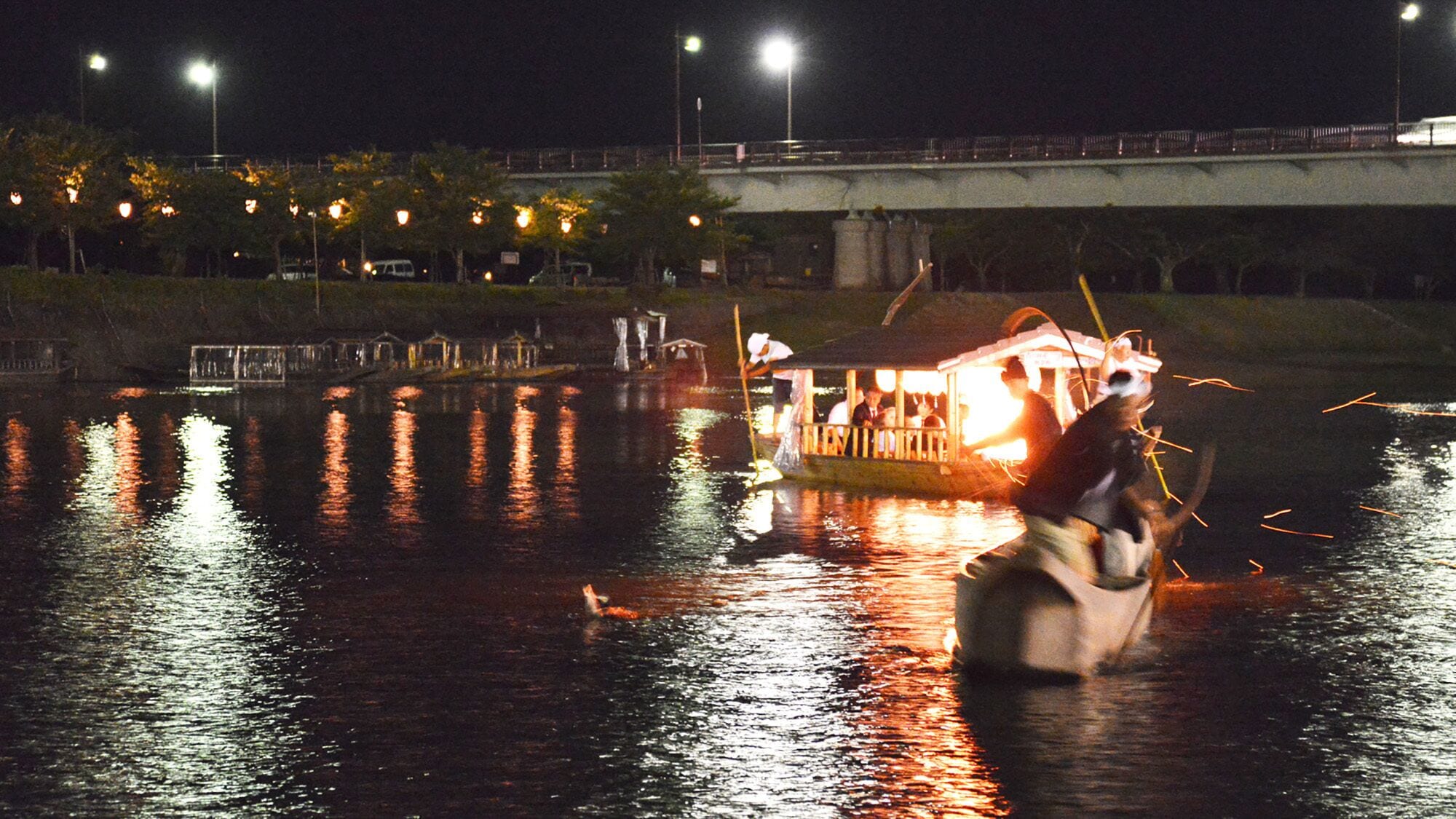 A nighttime scene with a bridge, illuminated buildings, and a boat on the water.