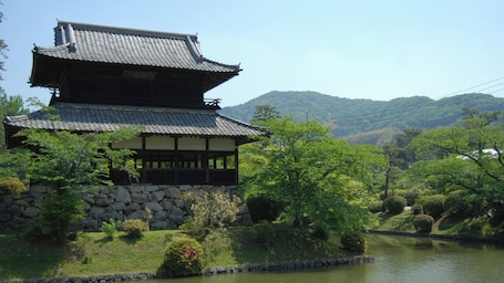 A traditional Japanese building with a tiled roof and wooden structure, surrounded by lush greenery and a body of water.