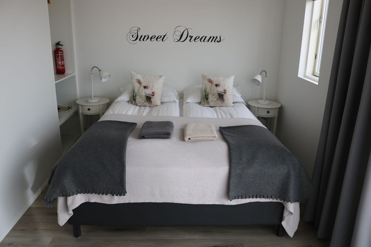 A neatly arranged bedroom with a bed, two bedside tables, a bedside lamp, a fire extinguisher, and a window with curtains.