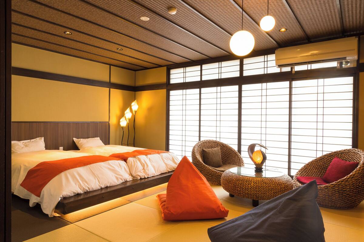 Una habitación de hotel con una cama, dos sillas de mimbre, una mesita y ventanas amplias.