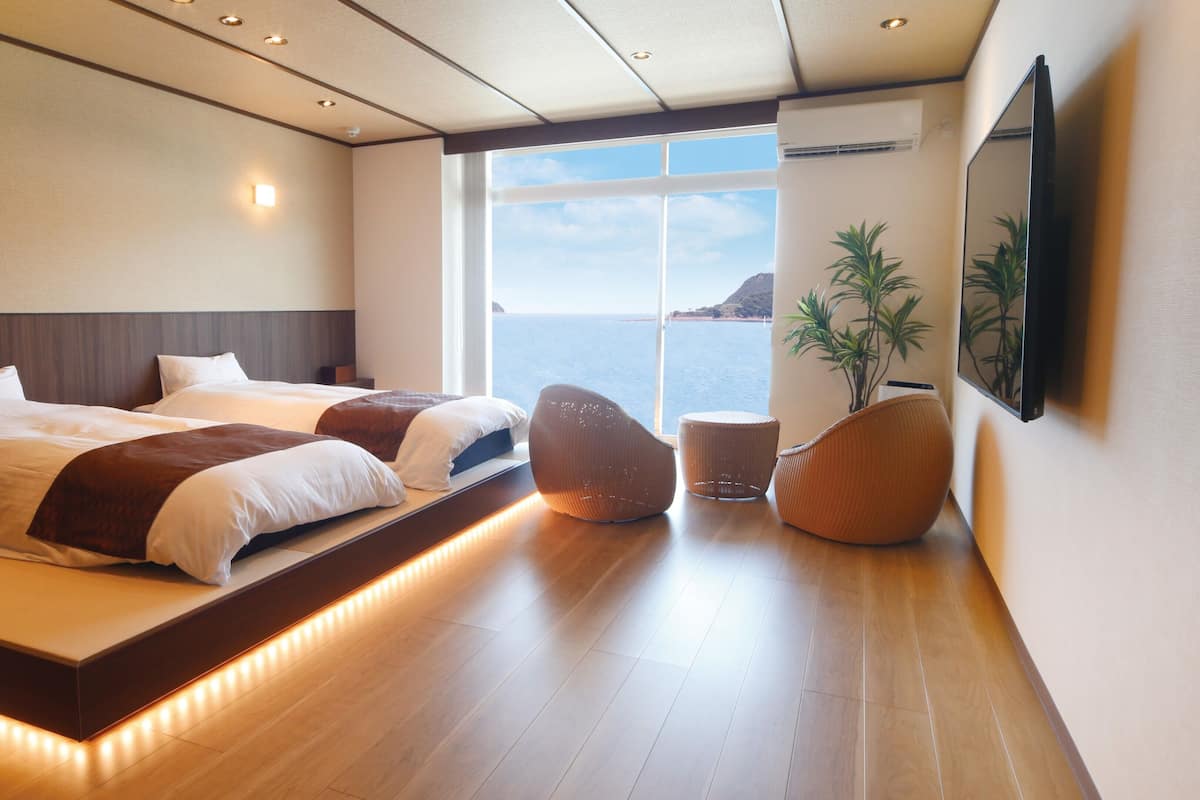 Una habitación de hotel moderna con una cama grande, una zona de estar con sillas, una mesita y un ventanal con vistas al mar.