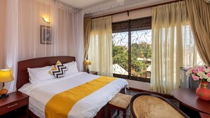 A hotel room with a large bed, a bedside table with a lamp, a chair, a desk with a vase of flowers, and a window with curtains. - Entebbe Travelle'rs Inn (Entebbe)