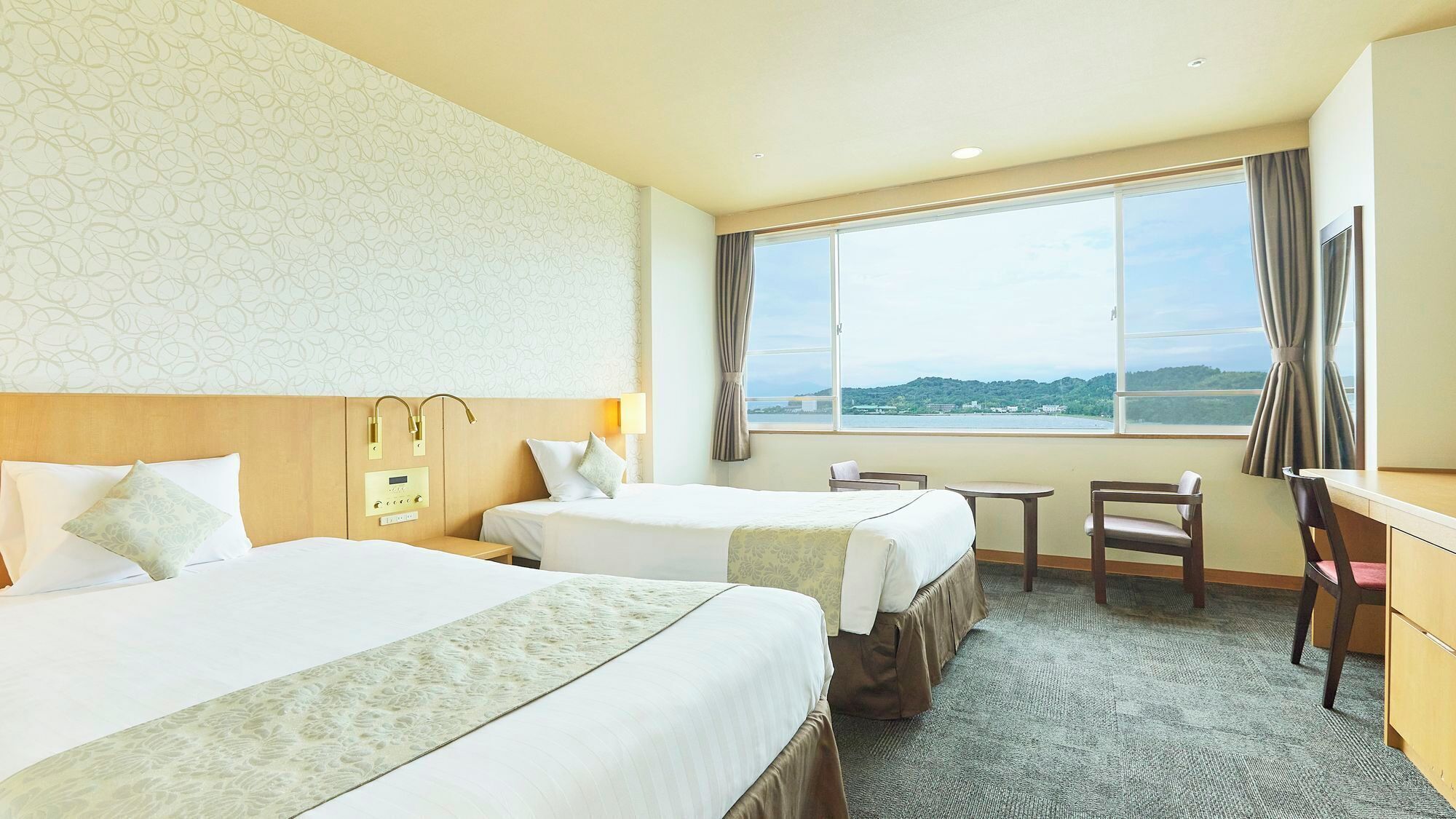 Une chambre d’hôtel avec deux lits, un bureau, une chaise, une petite table et une grande fenêtre donnant sur un beau paysage.