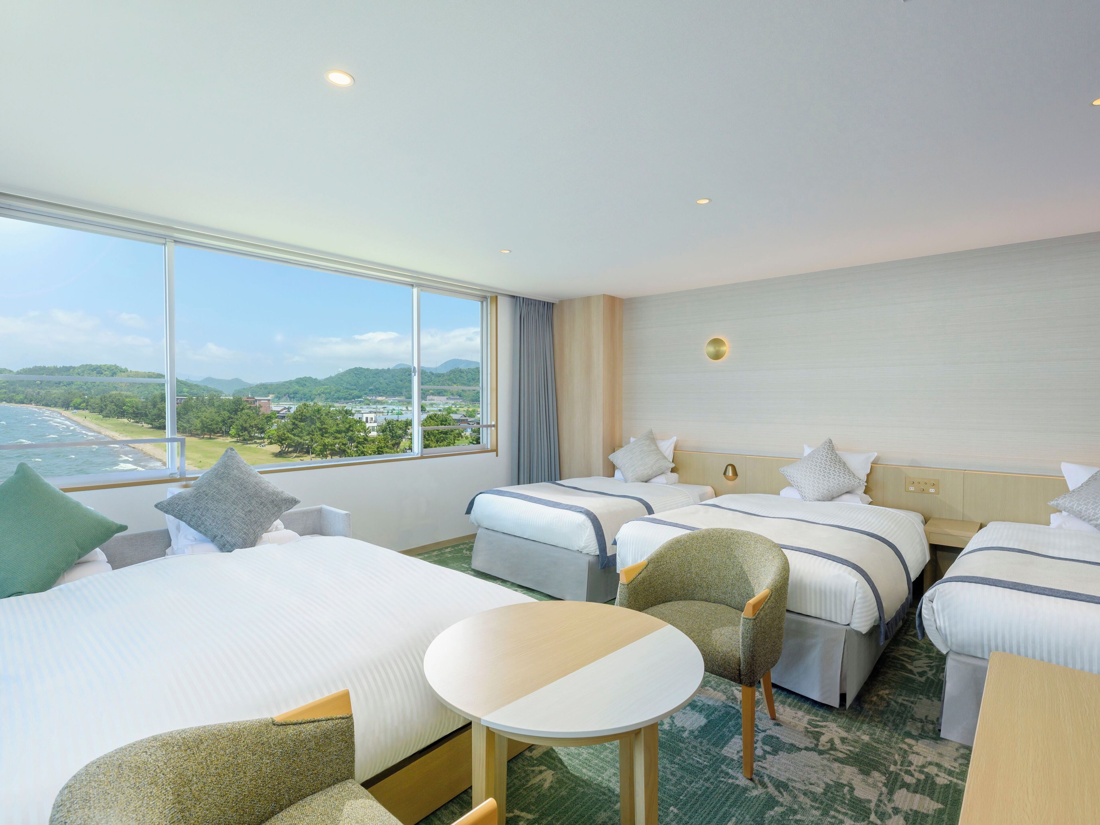 A hotel room with two beds, a small table, and a chair. There is a large window offering a view of the sea and greenery.