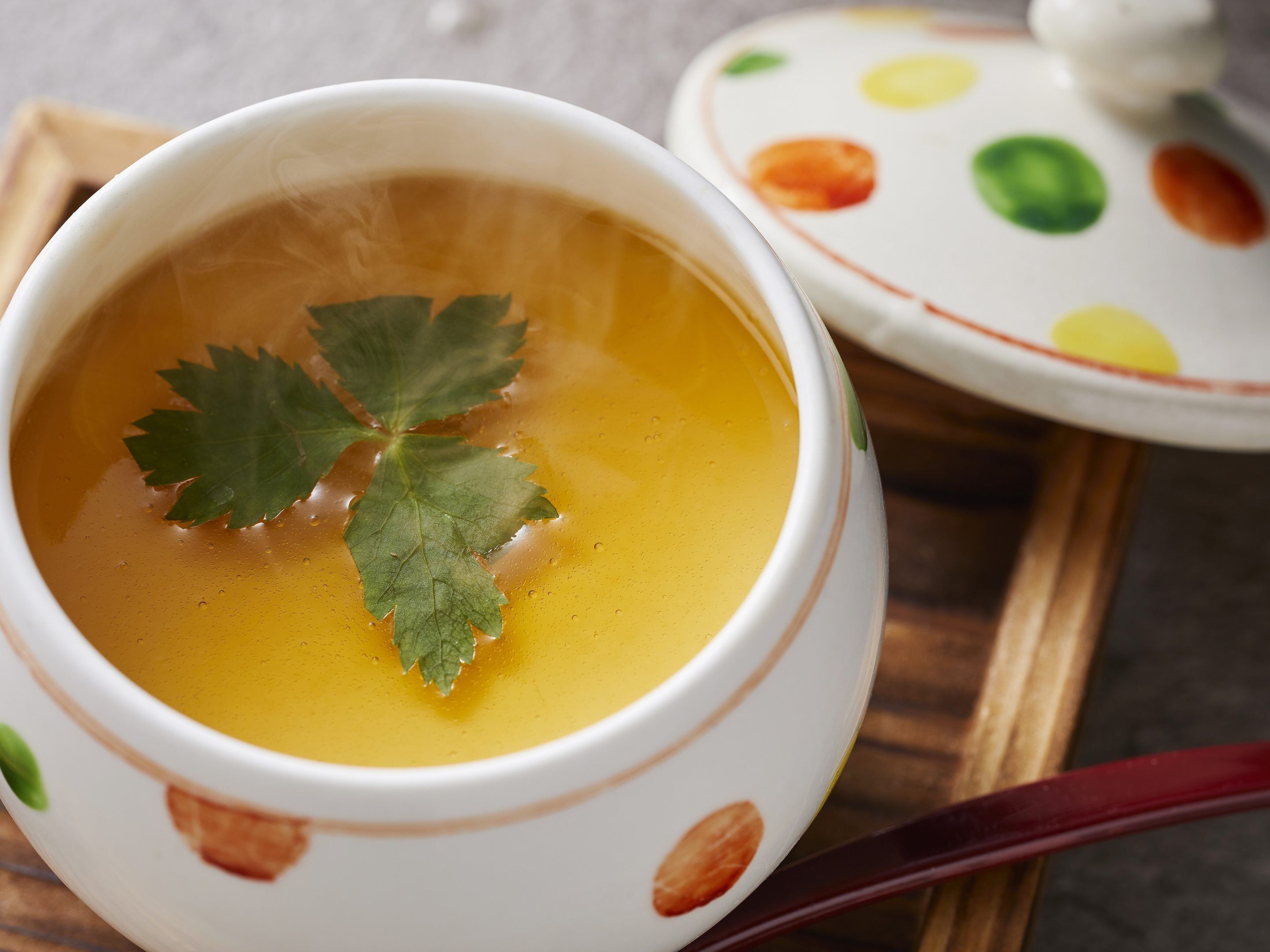 A bowl of soup with a leaf garnish, a spoon, and a patterned teapot on a tray.