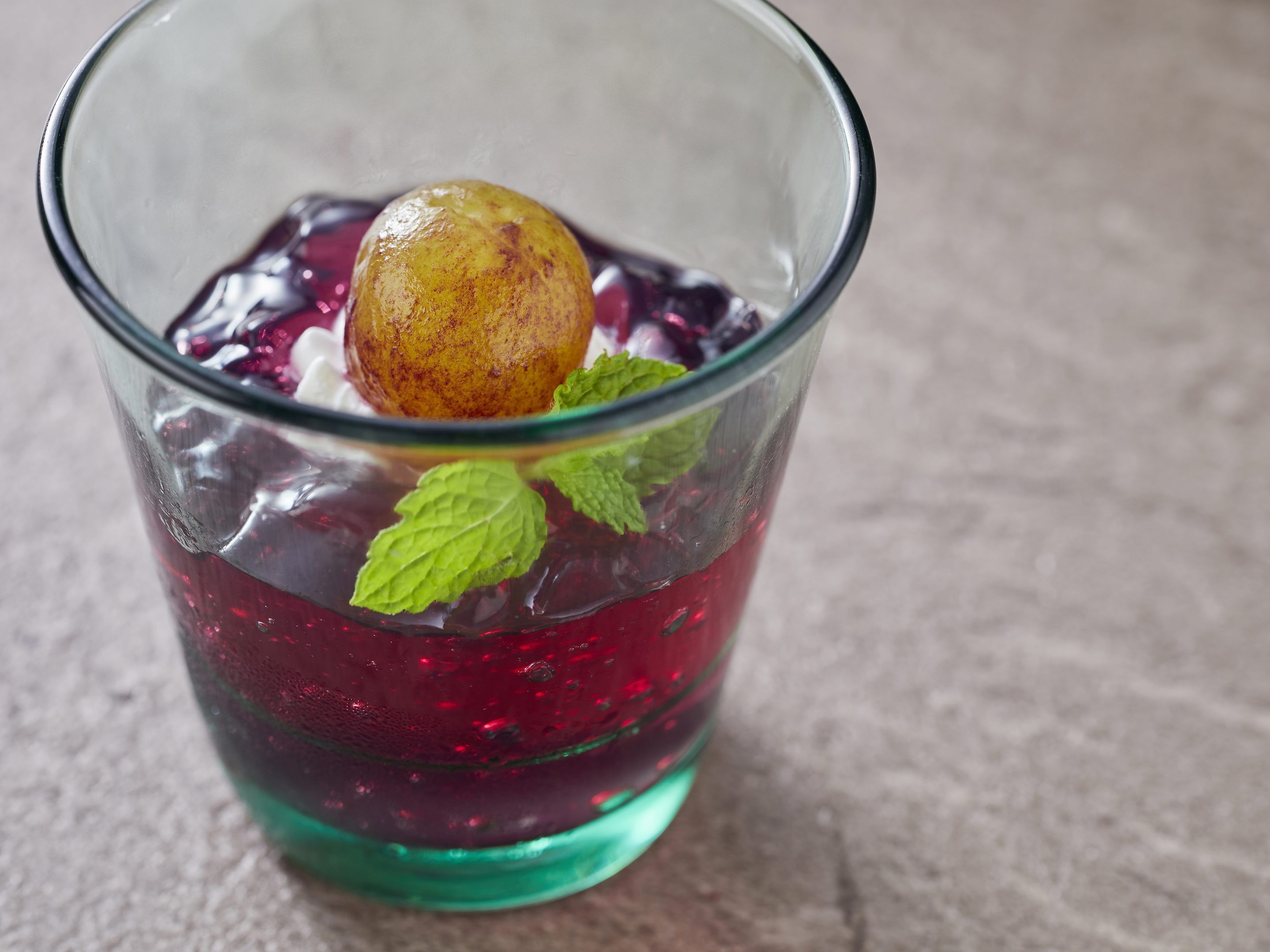 A glass with a red liquid, ice cubes, a fruit, and mint leaves.