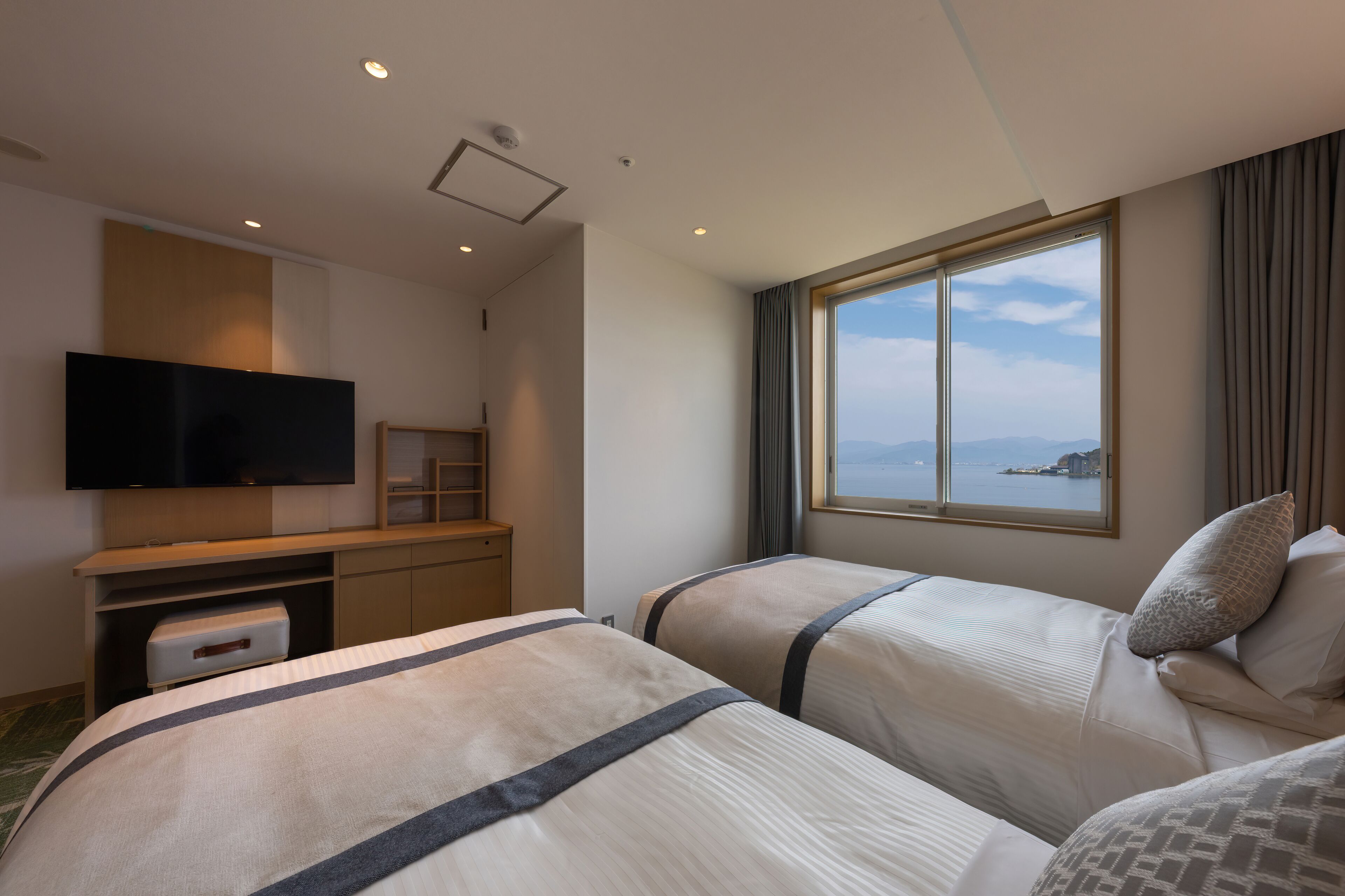 Habitación de hotel con dos camas, televisión, escritorio y ventana con vista al mar.