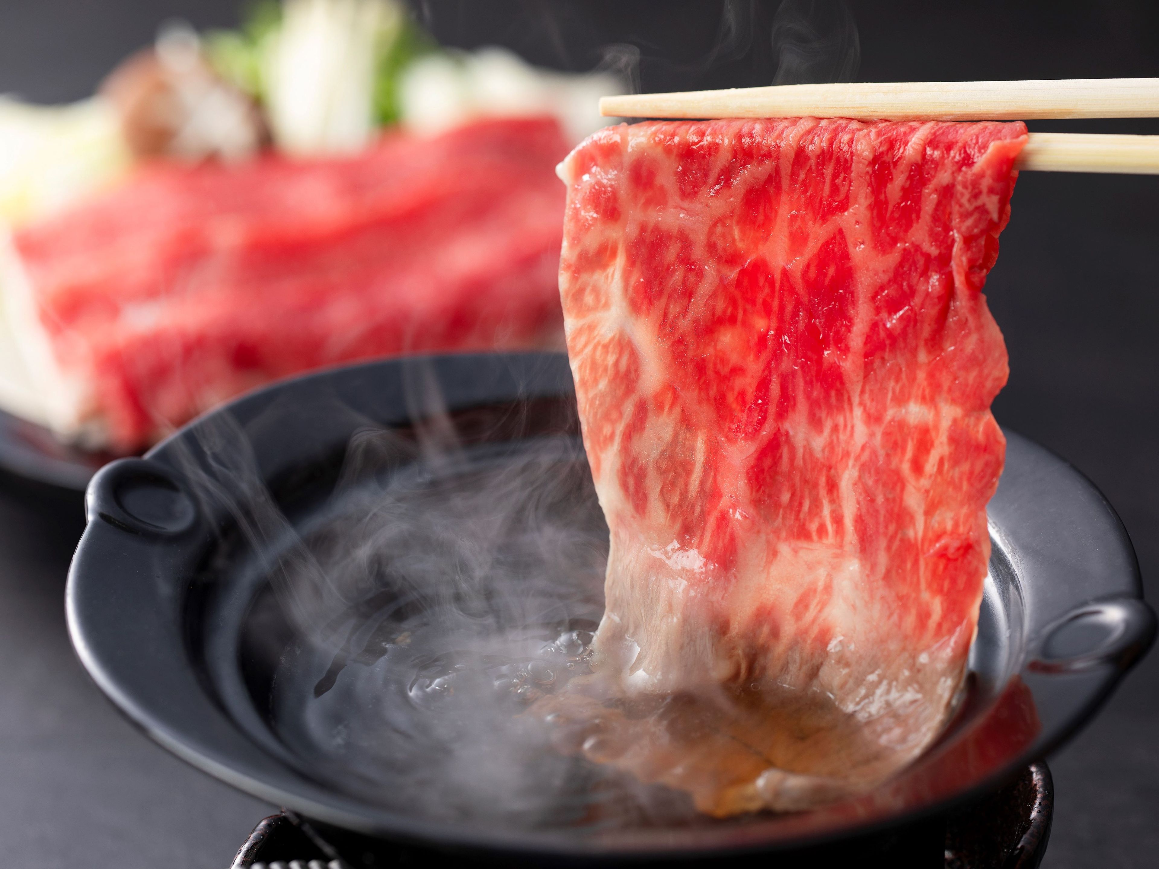 A close-up of a piece of raw meat being held by chopsticks over a steaming pot.