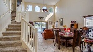 A spacious living area with a dining table set for a meal, a staircase with a ceiling fan, and a view of the outdoors through large windows. - Luxury 6BR / 3.5 Bathroom villa near Disney. Pool, Hot Tub, Games Room, etc. (Kissimmee)