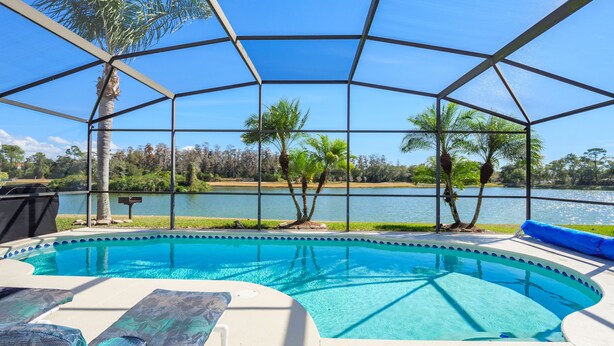 A pool with a clear view of a lake and trees, surrounded by a covered area with lounge chairs. - Luxury 6BR / 3.5 Bathroom villa near Disney. Pool, Hot Tub, Games Room, etc. (Kissimmee)