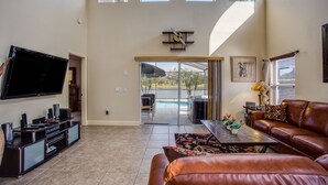A spacious living room with a flat-screen TV, leather sofa, and a dining area with a glass door leading to a pool. - Luxury 6BR / 3.5 Bathroom villa near Disney. Pool, Hot Tub, Games Room, etc. (Kissimmee)