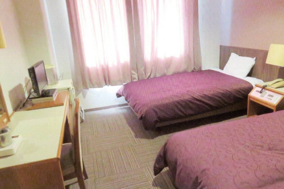 Habitación de hotel con dos camas, un escritorio, una silla, una lámpara y una ventana con cortinas.