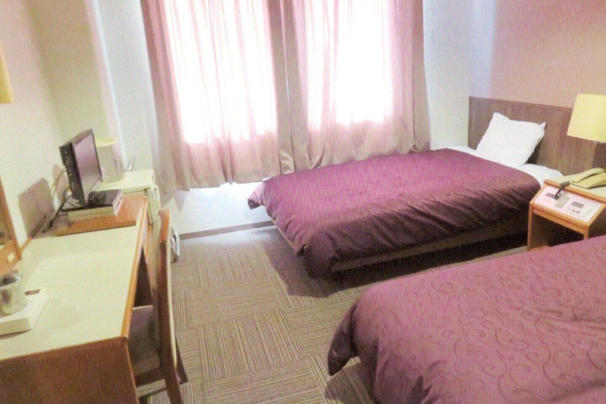 Habitación de hotel con dos camas, un escritorio, una silla, un ordenador y una mesita de noche con lámpara.