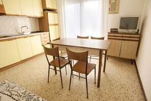 A compact kitchen with a dining table and chairs, a small TV on a cabinet, and a window with curtains.