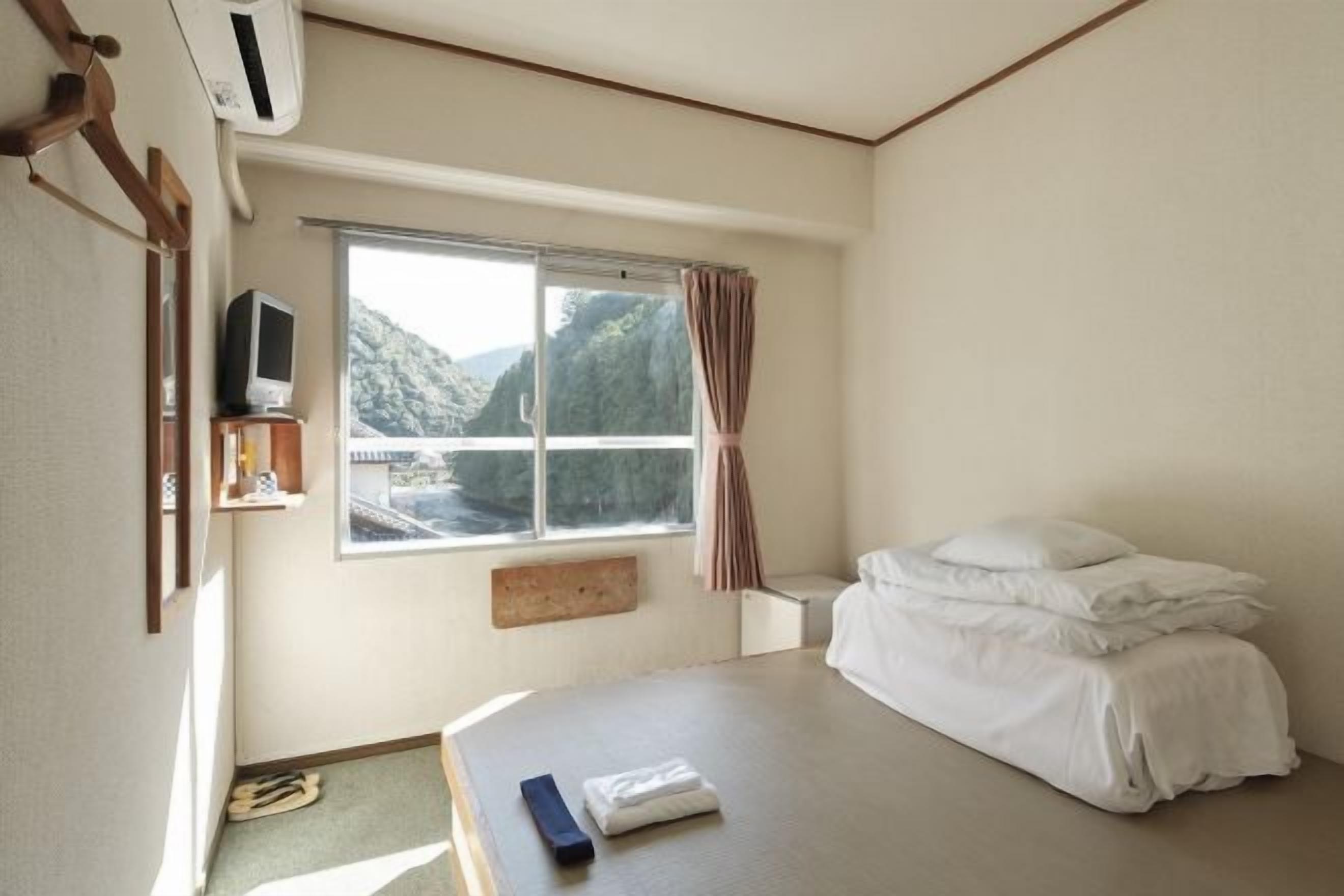 Een hotelkamer met een bed, een raam met uitzicht op een berg, een televisie op een plank en een afstandsbediening op het bed.