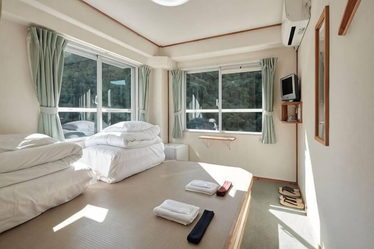 A hotel room with a bed, bedside table, TV, and windows with curtains.