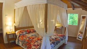 Een hemelbed met een bloemenbedekking, een nachtkastje met een lamp en een raam met uitzicht op groen.