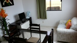 Een woonkamer met een eettafel, stoelen, een bank, een televisie, een ventilator en een raam met gordijnen.