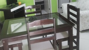 Een kamer met een eettafel met glazen blad, twee houten stoelen, een witte bank en een televisie op een zwart voetstuk. Er zijn twee deuren naar een kast en een badkamer.