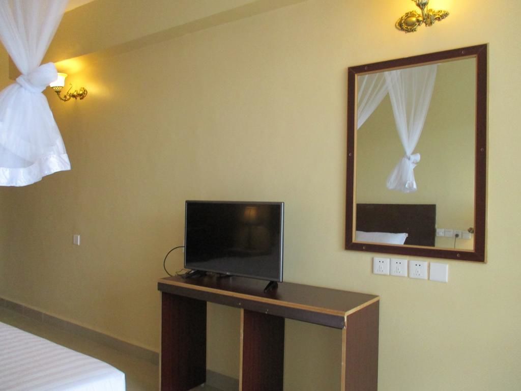 Habitación de hotel con cama, televisión sobre un soporte de madera, espejo fijado a la pared y aplique de luz.