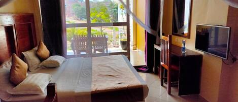 A hotel room with a large bed, a TV, a balcony with a view of the city, and a wooden desk.