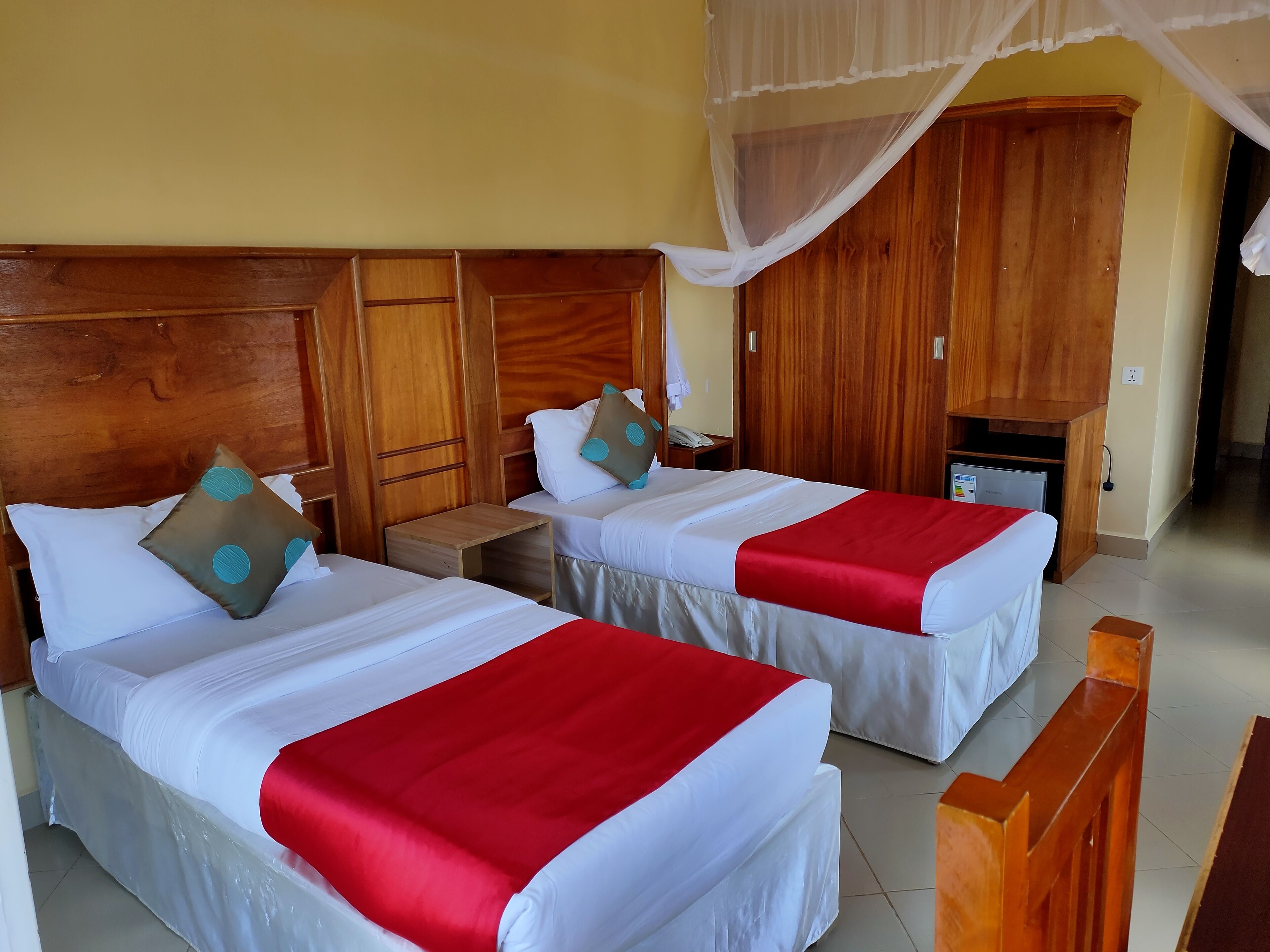 A hotel room with two beds, wooden headboards, a small bedside table, a nightstand, a wooden wardrobe, and a mosquito net.