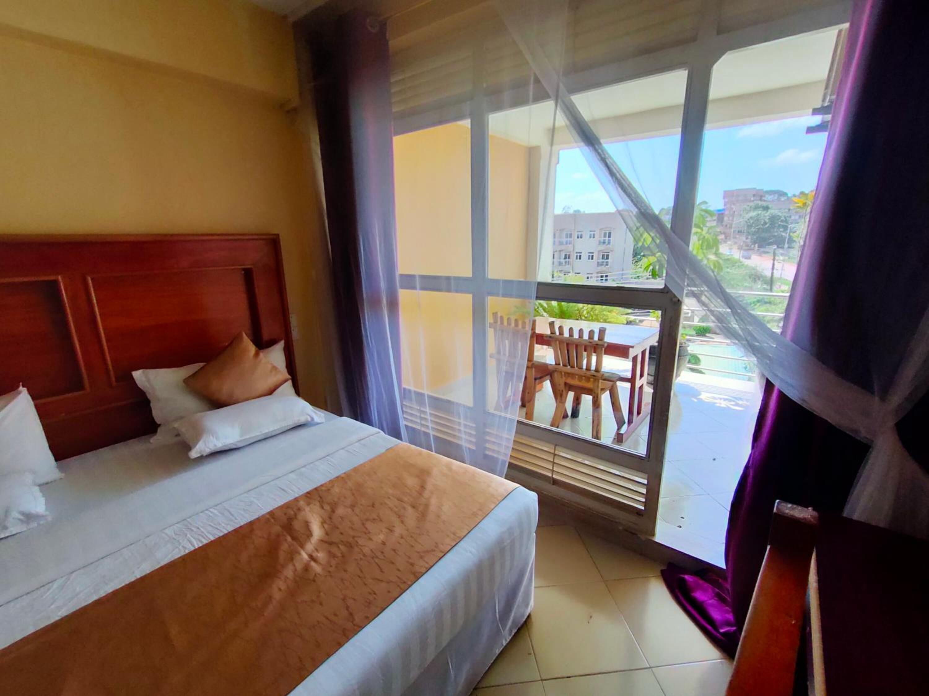 A hotel room with a large bed, a wooden headboard, a balcony with a table and chairs, and a view of buildings and greenery outside.