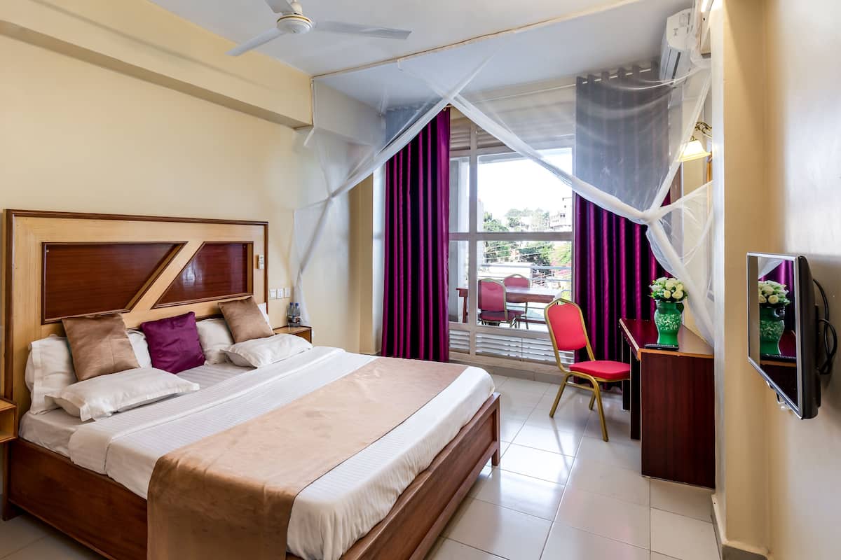 Quarto de hotel com uma cama grande, uma escrivaninha com cadeira, uma televisão e uma janela com cortinas.