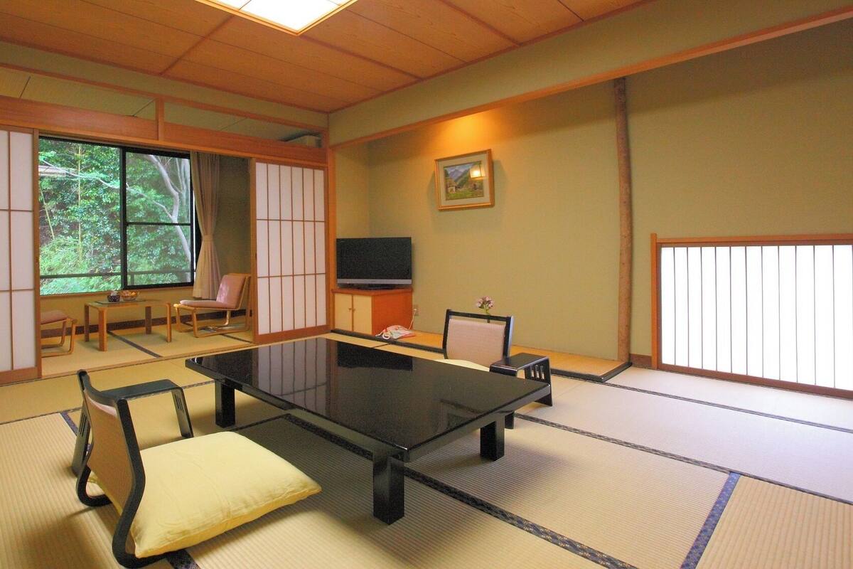 Una habitación tradicional japonesa con suelo de tatami, una mesa baja, sillas, un televisor y una ventana con vistas a la vegetación.