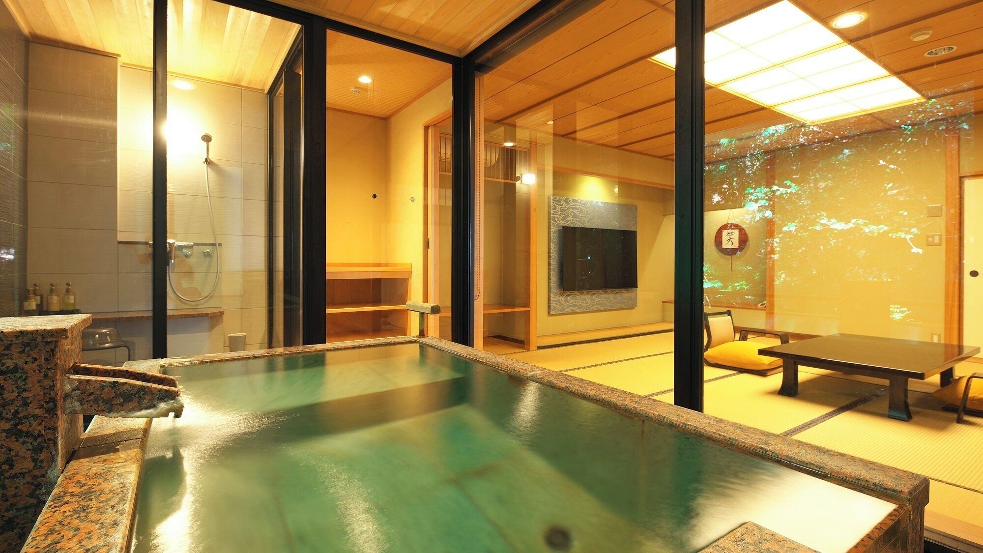 A traditional Japanese-style room with tatami flooring, a small indoor hot spring bath, and a sliding glass door leading to a larger space with a television and a wall-mounted screen.