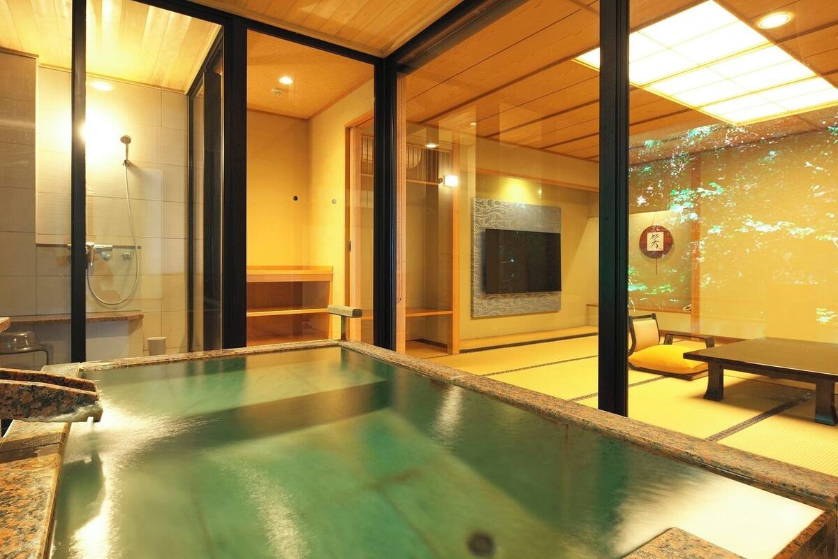 Una habitación tradicional japonesa con suelo de tatami, un pequeño baño de aguas termales interior y un puertas correderas de vidrio que dan a un espacio más amplio con un televisor y una pantalla montada en la pared.