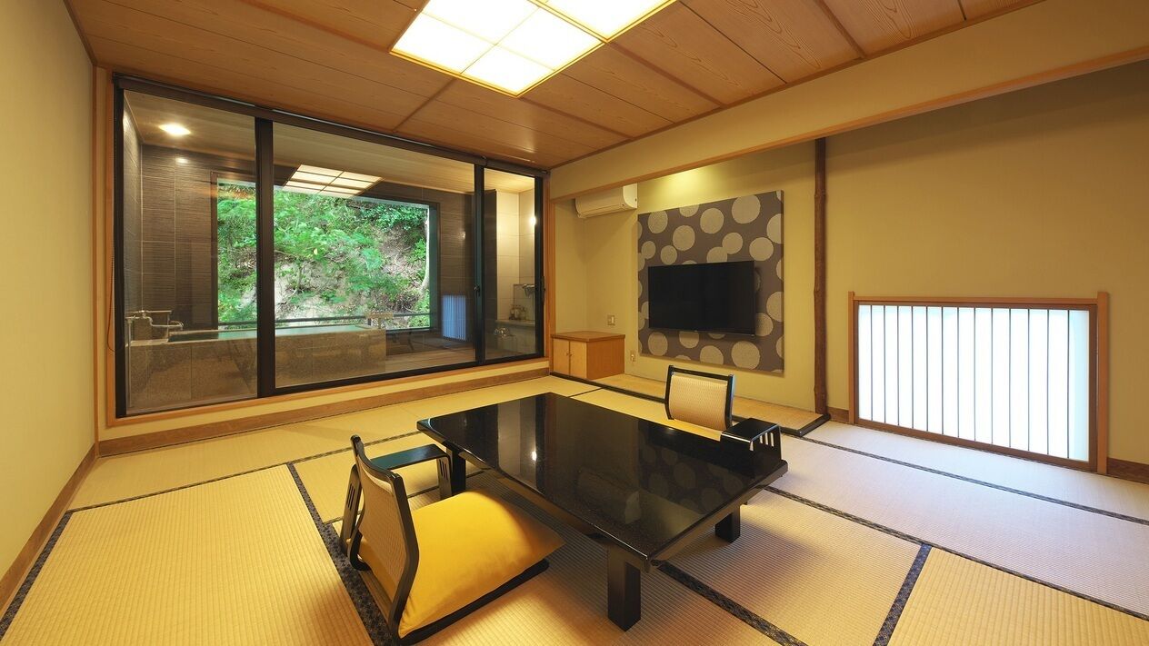 A traditional Japanese-style room with tatami flooring, a low table, a sofa, a sliding door with a view of greenery, and a mounted TV.