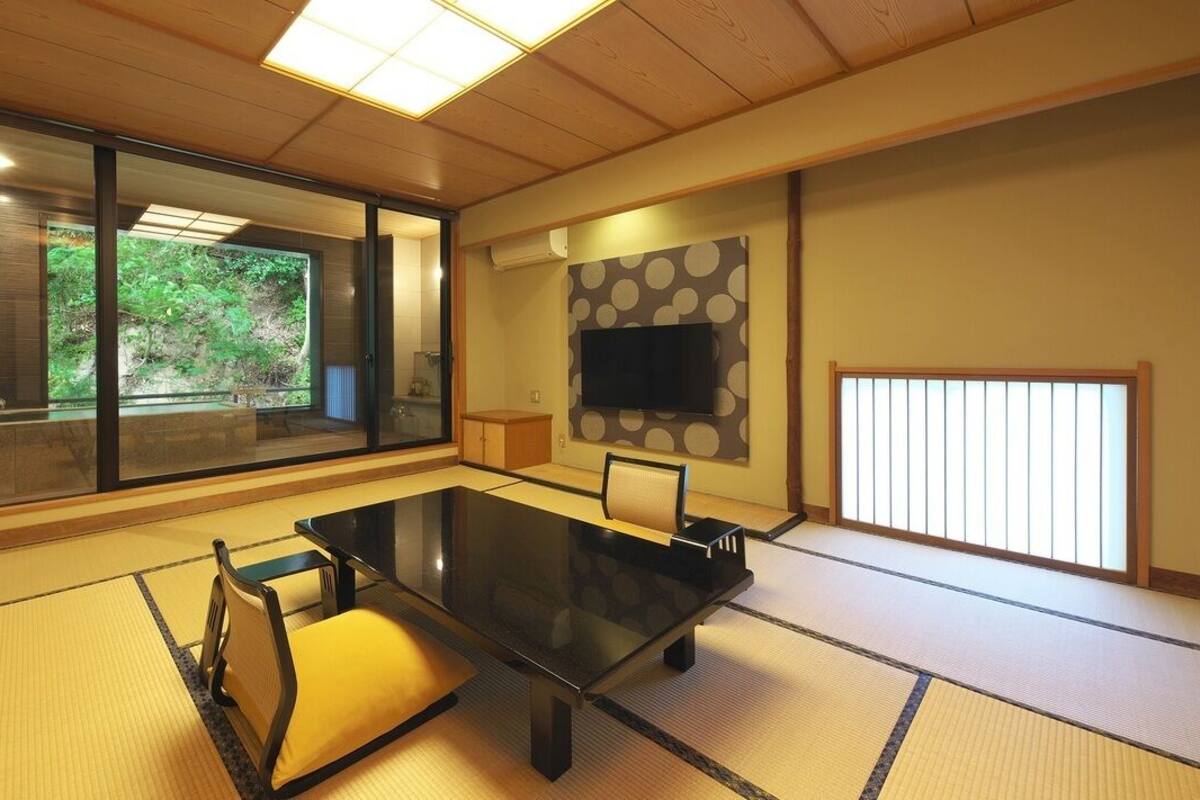 Una habitación tradicional japonesa con suelo de tatami, una mesita baja, un sofá, una puerta corredera con vistas a la vegetación y un televisor montado en la pared.