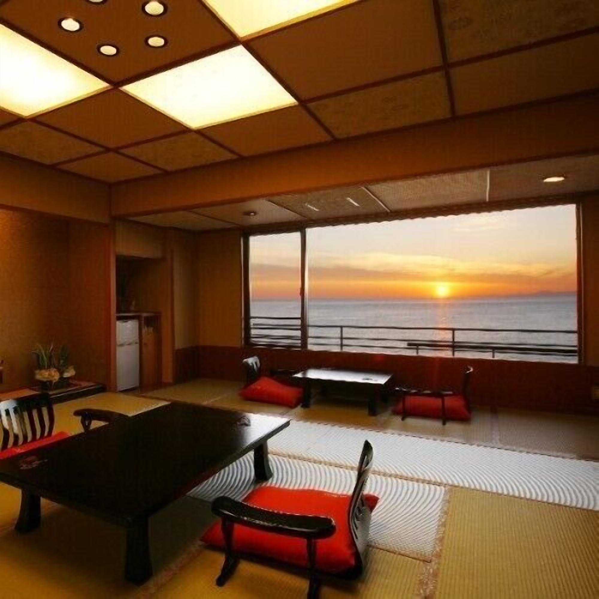 A room with a large window, a table, chairs, and a view of the sunset over the ocean.