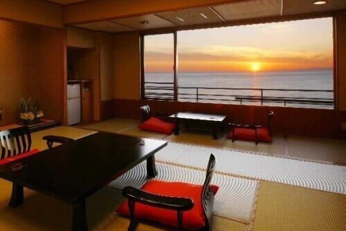 A room with a large window, a table, chairs, and a view of the sunset over the ocean.