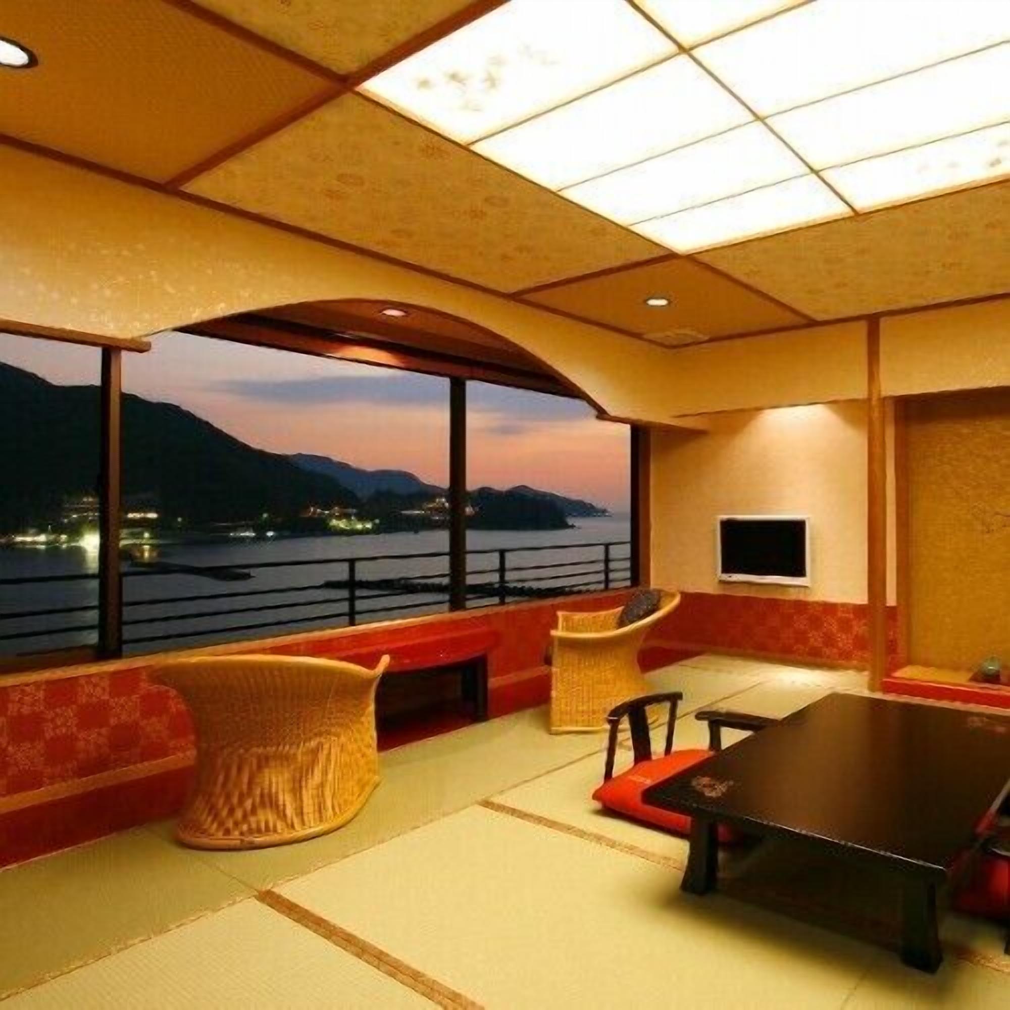 A room with a large window overlooking a body of water and mountains, a table, chairs, and a television.