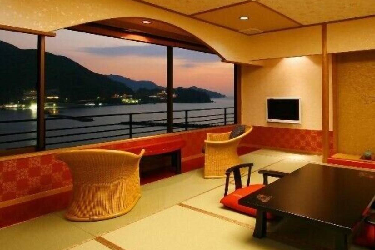 A room with a large window overlooking a body of water and mountains, a table, chairs, and a television.