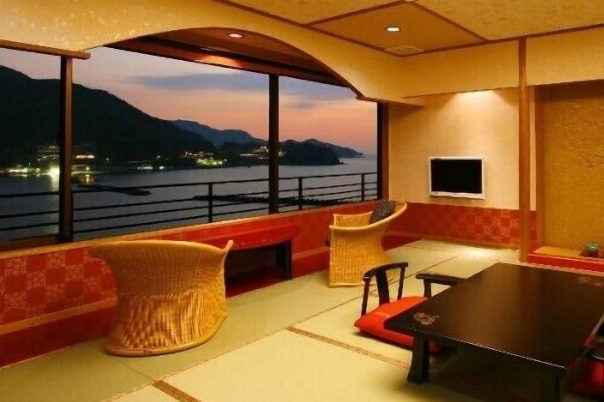 A room with a large window overlooking a body of water and mountains, a table, chairs, and a television.