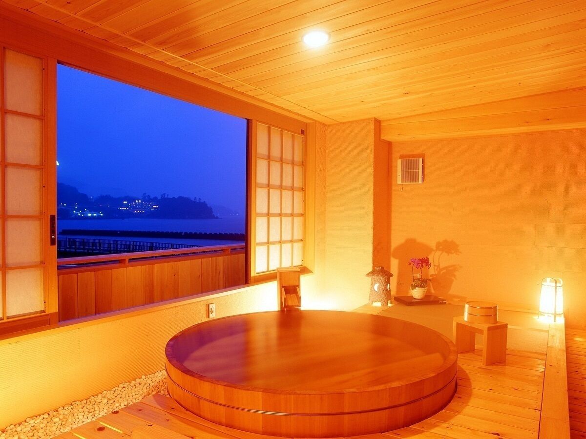 A wooden room with a large window, a wooden bathtub, and a small table with a potted plant.