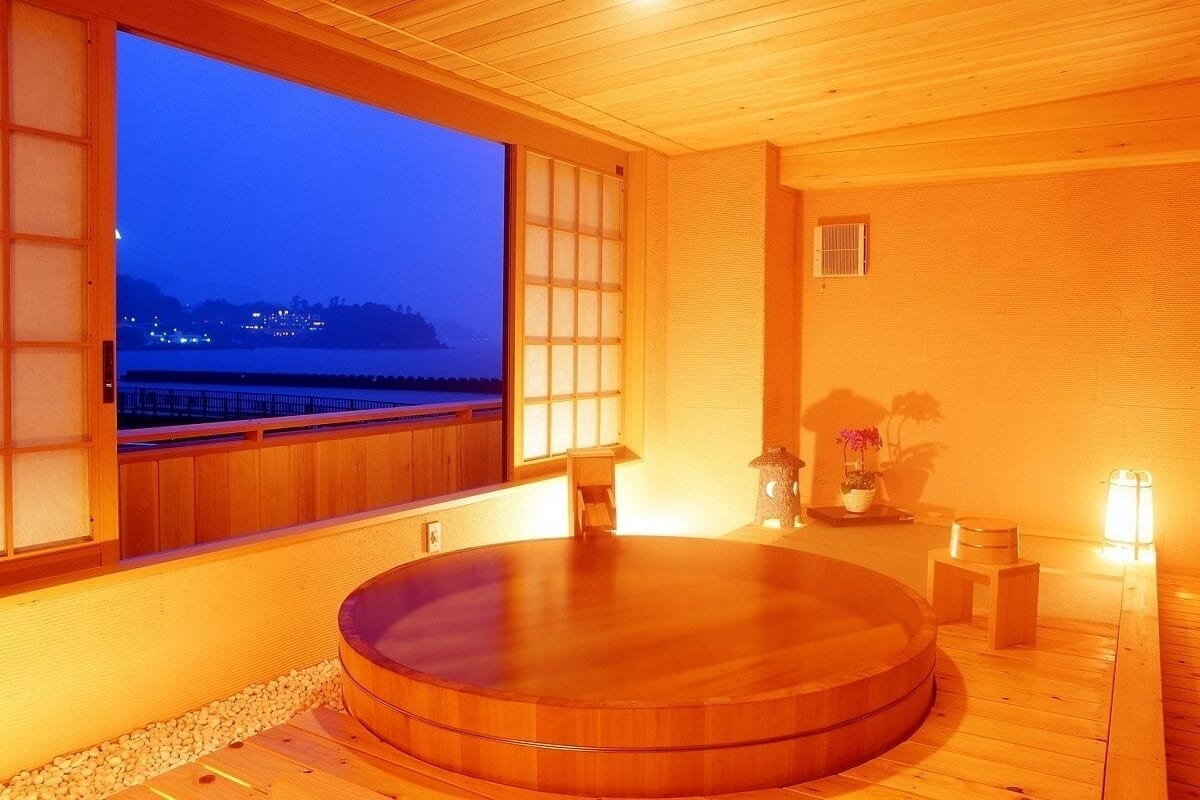 A wooden room with a large window, a wooden bathtub, and a small table with a potted plant.